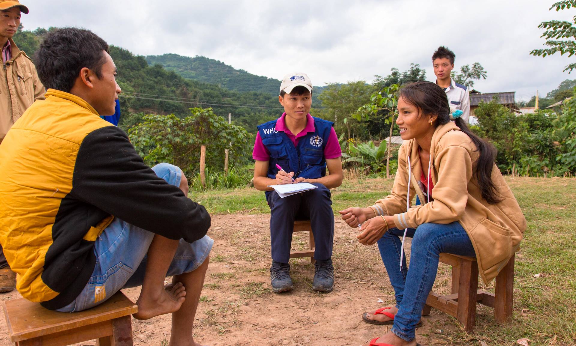 Three people sit on small wooden stools outside and talk